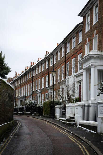 A row of traditional red-brick terraced houses with white window frames and decorative columns at the entrances, situated along a narrow, slightly curved street in a residential area. The scene features a small patch of snow on the pavement and some bushes and trees in front of the properties. A black lamppost stands near the sidewalk, and the houses have multiple stories with chimney stacks visible on the rooftops. The sky appears overcast, creating diffuse lighting. The street is clean, with no visible rubbish or debris, and the setting suggests a quiet suburban environment where private waste collection or on-site clearance might be undertaken by services such as House Clearance Twickenham, which specializes in rubbish removal within this area.