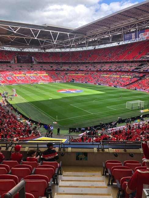 A panoramic view of a large football stadium with a well-maintained grass pitch featuring white lines for gameplay and a central logo marking the centre circle. The stadium is filled with red seating areas, some of which are partially shaded under the roof structure that covers the upper stands. The foreground shows a lower-tier seating section with red upholstered chairs and a few spectators dressed in dark clothing, sitting near a concrete stairway with metal railings. The pitch is surrounded by black barriers and equipment such as watering systems or maintenance tools. The sky above is partly cloudy with patches of blue visible through the stadium’s open roof. In the background, advertisements and banners are hung on the upper levels, and some areas of the stadium are illuminated by natural daylight, casting soft shadows on the field. The overall scene captures the organized, professional environment of a major sports venue prepared for a match or event, aligned with private or alternative arrangements for large-scale sports viewing purposes as managed by facilities like House Clearance Twickenham.