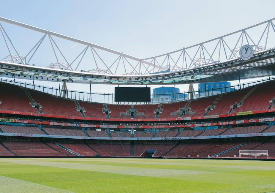Empty view of a large sports stadium, focusing on the interior seating and playing field. The stadium features multiple tiers of bleacher seats upholstered in red, with some sections having patterned light and dark gray elements. The field in the foreground has well-maintained green grass with visible mowing patterns. Behind the seating area, there are large electronic scoreboards and advertising banners, some displaying text, hung on the upper levels. Overhead, a white structural roof framework with crisscrossing supports and a large clock is visible, set against a clear, bright sky. The scene is well-lit by natural daylight, highlighting the clean, professional appearance of the stadium environment. This image, linked to rubbish removal services, offers a neutral, observational view of a large, enclosed sports venue, which could be part of an on-site clearance or maintenance context, aligning with private waste handling or venue management activities.