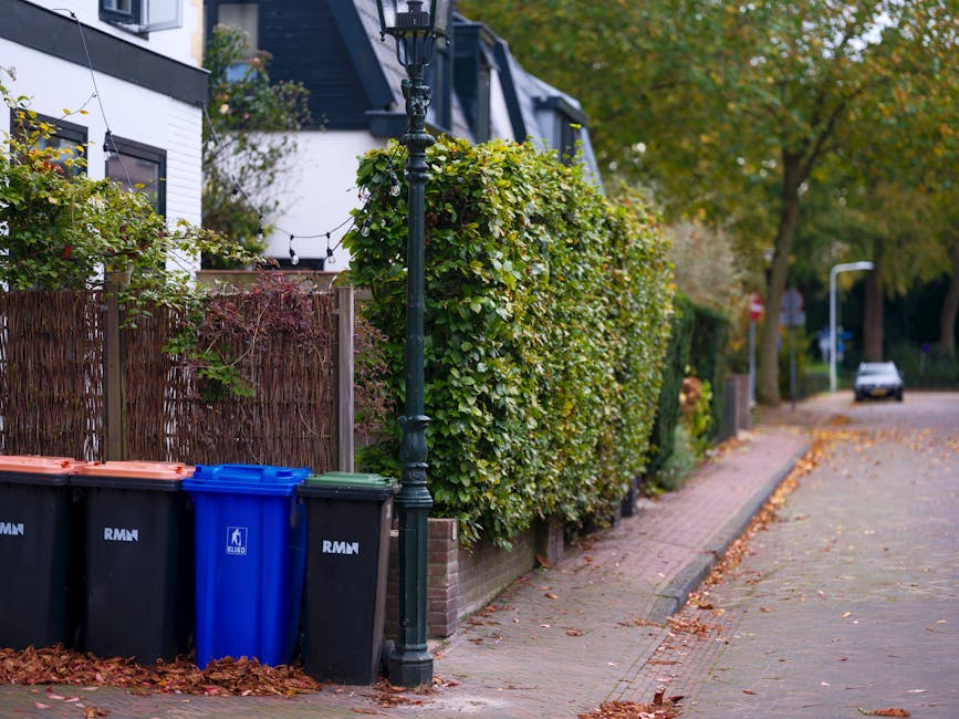 In the foreground, four wheeled rubbish bins are positioned along the edge of a residential sidewalk; the first three bins are black with orange lids, while the fourth is blue with a matching lid, all made of plastic with a smooth surface. Behind the bins, there is a wooden fence with a brown, woven texture, partially obscured by dense, green foliage from a nearby shrub or small tree. A vintage-style black street lamp with decorative detailing stands adjacent to the bins, casting ambient light over the scene. The sidewalk is paved with grey concrete slabs, and fallen autumn leaves are scattered across its surface, leading towards a quiet street with a single parked car visible in the distance. On the far side of the street, there are additional lampposts and trees with green and yellow leaves indicating a seasonal transition. The environment appears calm and well-maintained, suitable for independent waste management services such as rubbish collection or on-site clearance, consistent with services offered by House Clearance Twickenham for properties with narrow access.