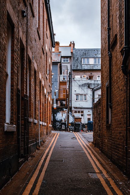 A narrow alleyway situated between two brick buildings with reddish-brown facades, featuring multiple vertical drainpipes and windows with white frames. The paved ground has double yellow lines along both edges, indicating parking restrictions. At the alley's end, there is a pile of mixed waste materials including large black bins and discarded packaging, which appears to be part of a rubbish collection or clearance process. Behind the waste, a small wooden staircase attached to a grey concrete building leads upward, and additional structures with flat and pitched roofs, some with chimneys, are visible in the background under an overcast sky. The scene, associated with independent waste disposal methods, aligns with the context of rubbish removal services in a residential or urban setting, with the appearance of an on-site clearance operation by House Clearance Twickenham.